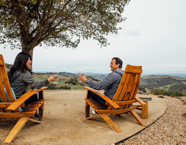 A couple sit outside and relax with to drink wine while overlooking a hill