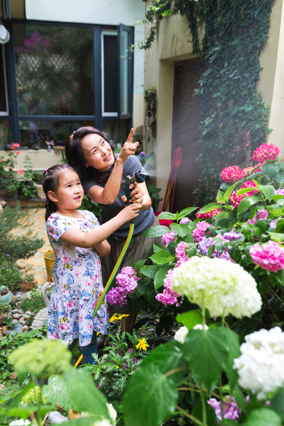 An older woman and child stand in a garden holding a hose surrounded by summer flowers.