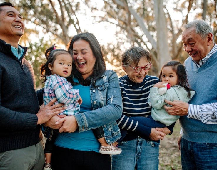 Parents and grandparents holding their grandchildren and laughing outdoors with sunlight trees in the background.